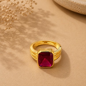 Può includere: Un anello color oro con una grande pietra preziosa rettangolare rosso scuro incastonata in una montatura liscia. La fascia è composta da tre fili dorati sottili e paralleli che convergono alla base dell'incastonatura, creando un aspetto a strati.