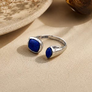 May include: A silver-toned ring with two dark blue, oval-shaped stones. The ring has an open design, with one stone at each end. The ring is set against a neutral background.
