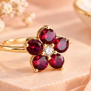 May include: A gold ring featuring a flower-shaped setting with five round, deep red gemstones as petals and a single, small, clear, round-cut diamond at the centre. The band is polished yellow gold, and the stones are held in place by gold prongs.