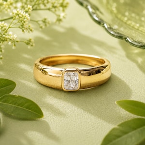 Könnte beinhalten: Ein goldener Bandring mit einem einzelnen, rechteckig geschliffenen, klaren Edelstein in einer Zargenfassung. Der Ring hat eine glatte, polierte Oberfläche und ein dickes, abgerundetes Profil, das ein schlichtes Design mit einem zentralen Stein zeigt.