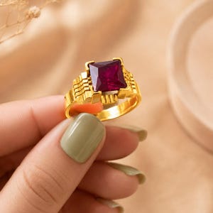 Op de afbeelding: Een gouden ring met een grote, vierkant geslepen, diep magentakleurige edelsteen in een gestructureerde, getrapte metalen band. De zetting heeft een geometrisch, getrapt ontwerp aan beide zijden van de centrale steen voor een robuuste look.