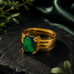 Puede incluir: Un anillo de oro con una gran gema verde de talla esmeralda. El anillo tiene una banda gruesa de oro y está sobre una superficie oscura y texturizada. Hay follaje verde en el fondo.