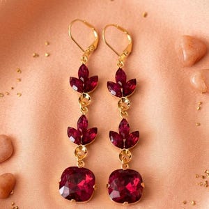 May include: A pair of gold-tone drop earrings featuring two stacked leaf-shaped deep red crystals and a single square-cut deep red crystal at the base. The earrings are suspended from gold-tone lever-back closures, showcasing a faceted design.