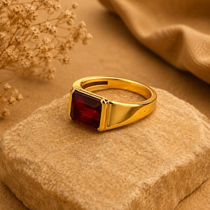 May include: A gold ring featuring a large, rectangular, deep red gemstone set in a smooth, polished metal band. The ring has a substantial, solid design with a flat top setting that holds the dark red stone securely in a bezel-style frame.