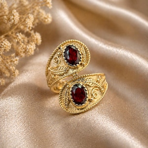 Peut inclure: Une bague dorée ornée de deux pierres précieuses ovales rouge foncé. La bague présente un design ouvert avec des détails ornés, notamment des motifs de feuilles et de volutes. La bague est présentée sur une surface dorée satinée.