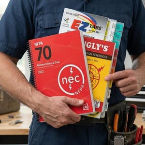 May include: A person holding a stack of electrical code books. The red book reads "NFPA 70 National Electrical Code 2023". Other books include "EZ Tabs for the NEC" and "Ugly's Electrical References". The books are color-coded and have tabs.