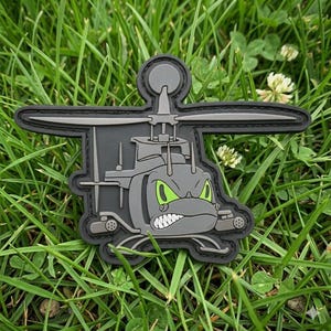 May include: A gray tactical morale patch shaped like a helicopter with a cartoonish, aggressive face. The helicopter features bright green eyes, a bared-teeth grin, and a top rotor blade. The design is rendered in a flat, molded rubber style.