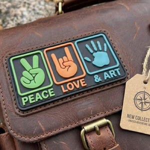 Può includere: Una borsa in pelle marrone con una toppa rettangolare che presenta tre icone: un segno di pace verde, un gesto della mano rock arancione e un'impronta di mano azzurra. Sotto le icone, il testo recita PEACE LOVE & ART. È attaccata un'etichetta beige.
