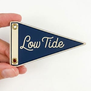 May include: A triangular navy blue pennant featuring the text Low Tide in a cream-colored script font. The pennant has a cream-colored vertical border on the left side with two brass-colored metal grommets for hanging or attachment purposes.