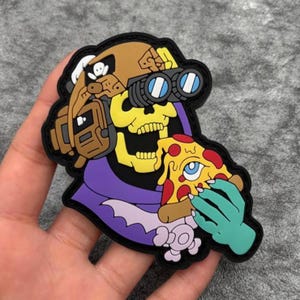 May include: A rubberised patch featuring a yellow skull character wearing a tan tactical helmet with night vision goggles. The character has a purple hood and is holding a slice of pizza with a blue eye in the centre. The design has a black border.