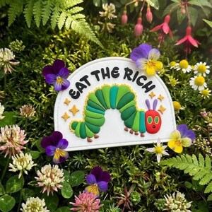 May include: A semi-circular white patch featuring a green caterpillar with a red head and purple antennae. The text EAT THE RICH is printed in black capital letters above the caterpillar, surrounded by small yellow four-pointed star shapes.