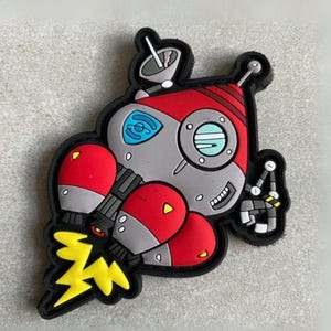 May include: A rubberized shoe charm shaped like a cartoon rocket ship in red, gray, and black. The design features a yellow lightning bolt exhaust, a satellite dish, a blue window, and a mechanical claw arm extending from the side of the craft.