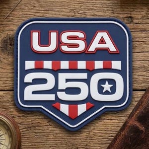 May include: A shield-shaped patch featuring a dark blue background with white and red accents. The text USA appears in white with red outlines above the number 250, which is also in white. A small white star is integrated into the zero.