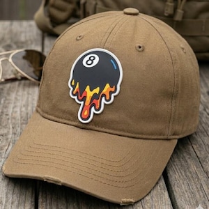 May include: A tan baseball cap featuring a centered patch of a black billiard ball with the number eight in white. The bottom of the ball design appears to be melting in shades of yellow, orange, and red with a white outline on the fabric.