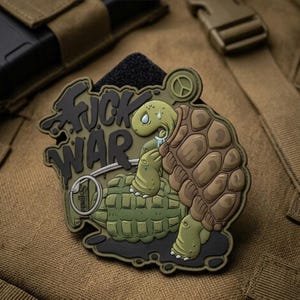 May include: A rubberised morale patch featuring a cartoon turtle leaning on a green hand grenade. The text FUCK WAR is written in black graffiti-style lettering. The turtle has a brown shell and light green skin, with a peace symbol icon above.
