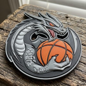 May include: A circular patch featuring a stylized gray dragon with sharp teeth, red eyes, and a dark red tongue, clutching a textured orange basketball with its clawed hand. The design is rendered in a layered, three-dimensional relief style.