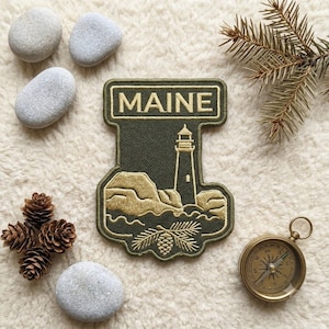 May include: An olive green embroidered patch featuring the word MAINE in a rectangular border at the top, with a lighthouse, rocky coastline, and pine branch design below. The patch has a dark green stitched edge and a textured fabric surface.