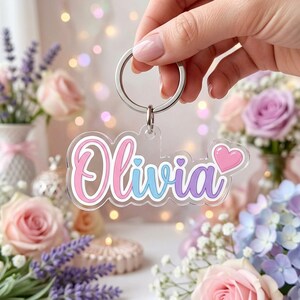 May include: A clear acrylic keychain featuring the name Olivia in a stylized script font. The letters transition from pink to blue to purple, with a small pink heart icon at the end. The keychain is attached to a silver metal split ring.