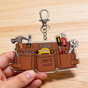 May include: A brown tool belt keychain with various tools, including a hammer, screwdriver, and wrench. The keychain has a silver clasp and the name "JACK 2025" printed on the front. The tools are arranged in pockets.
