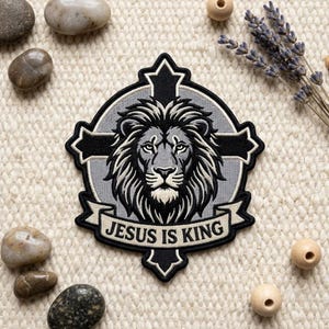 Könnte beinhalten: Ein kreisförmiger bestickter Aufnäher mit einem schwarz-grauen Löwenkopf, der über einem schwarzen Kreuz zentriert ist. Unter dem Löwen befindet sich ein Banner mit dem Text "JESUS IS KING" in schwarzer Schrift. Der Aufnäher hat einen dicken schwarzen Rand.