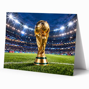 Op de afbeelding: Een wenskaart met de FIFA World Cup-trofee op een groen veld. De gouden trofee staat op de voorgrond, met op de achtergrond een stadion vol toeschouwers en heldere lichten. De kaart is wit.