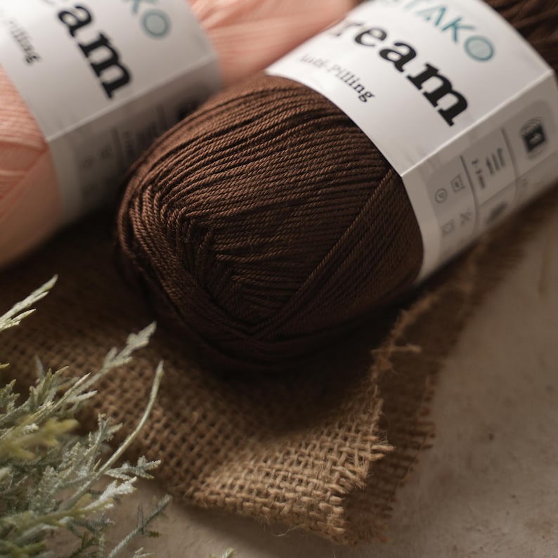 May include: Close-up of two yarn skeins. One is a rich brown color, and the other is a light peach. The yarn is labeled with the word "Dream" in a bold font. The skeins are resting on a burlap surface, with some greenery in the foreground.