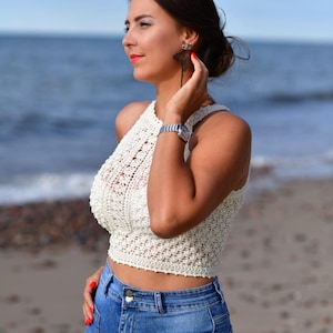 May include: A cream-colored, halter-style crochet top paired with blue denim shorts. The top features an open, textured weave. The model is wearing a silver watch and dragonfly earrings. The background shows the ocean and a sandy beach.