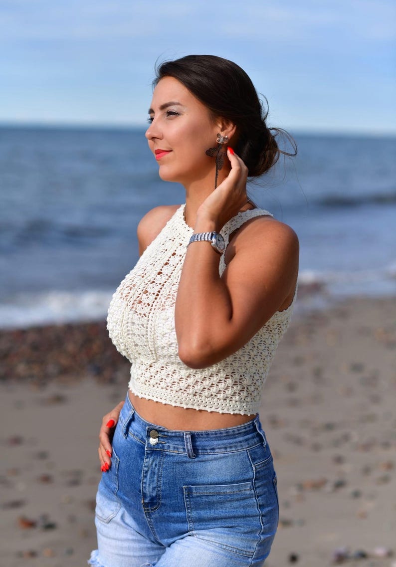 May include: A cream-colored, halter-style crochet top paired with blue denim shorts. The top features an open, textured weave. The model is wearing a silver watch and dragonfly earrings. The background shows the ocean and a sandy beach.