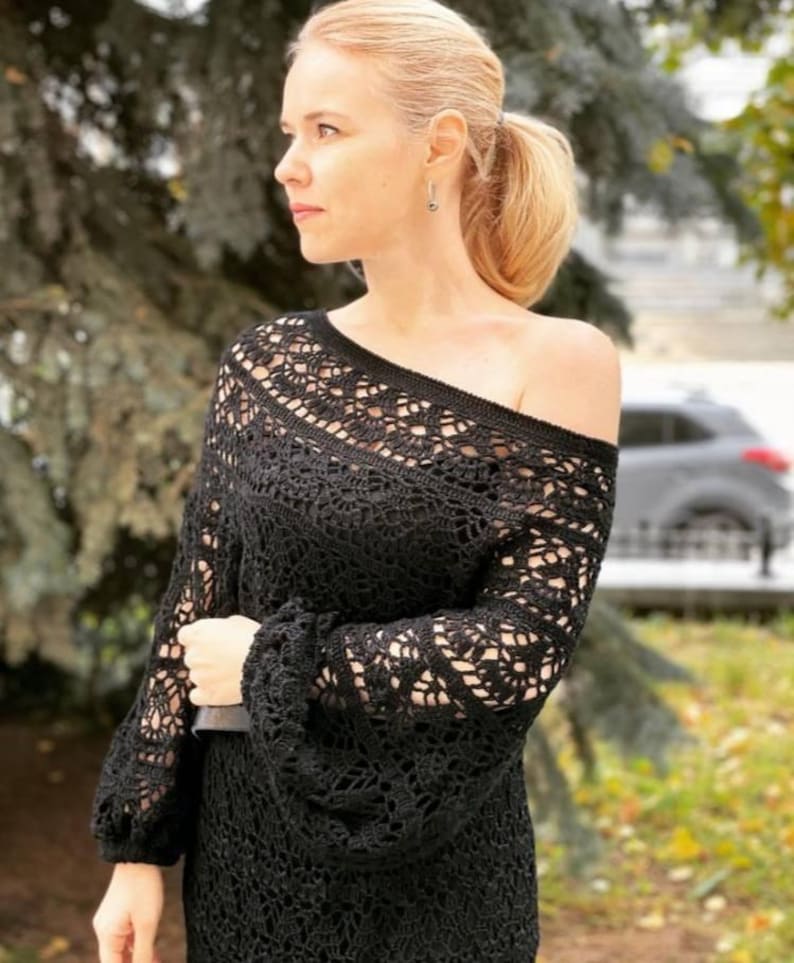 May include: Black crochet off-the-shoulder top with long sleeves. The top features an open, lacy design and a fitted waist. The sleeves have a slight puff at the shoulder and are fitted at the wrist. The top is worn outdoors.