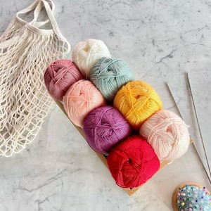 May include: A wooden tray holds nine colorful yarn balls in shades of pink, purple, yellow, green, and red. A white mesh bag and knitting needles are also present. The yarn balls are arranged in a diagonal row, showcasing the variety of colors.