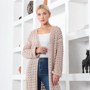 May include: A long, open-front, beige crochet cardigan. The cardigan has a textured, open weave design and long sleeves. The model is wearing the cardigan over a white top and blue jeans. The background features white shelves with decorative items.