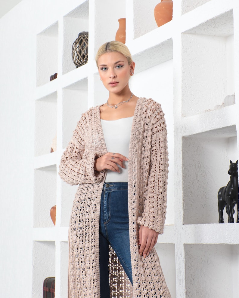 May include: A long, open-front, beige crochet cardigan. The cardigan has a textured, open weave design and long sleeves. The model is wearing the cardigan over a white top and blue jeans. The background features white shelves with decorative items.