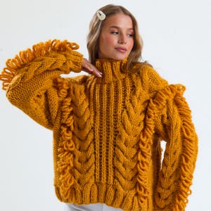 Pode incluir: Camisola de malha canelada amarelo mostarda com gola alta e detalhes de franjas nas mangas. A camisola apresenta um design de malha grossa e texturizada. As mangas t&ecirc;m um detalhe de franjas &uacute;nico, adicionando um toque elegante &agrave; pe&ccedil;a.