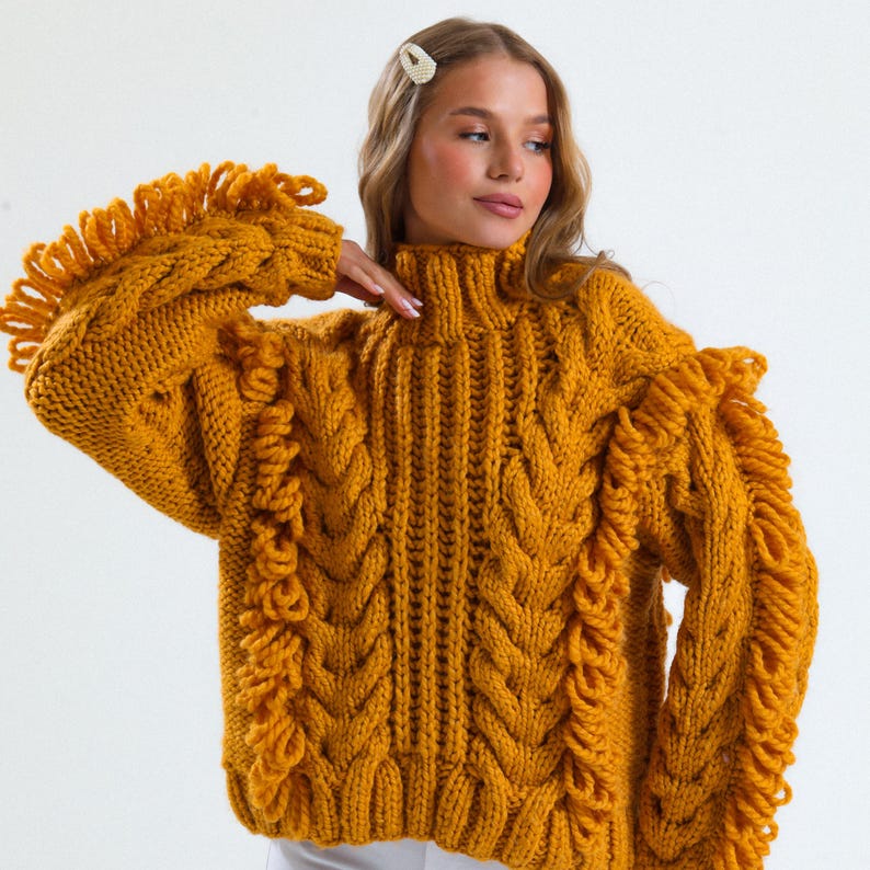 Pode incluir: Camisola de malha canelada amarelo mostarda com gola alta e detalhes de franjas nas mangas. A camisola apresenta um design de malha grossa e texturizada. As mangas t&ecirc;m um detalhe de franjas &uacute;nico, adicionando um toque elegante &agrave; pe&ccedil;a.