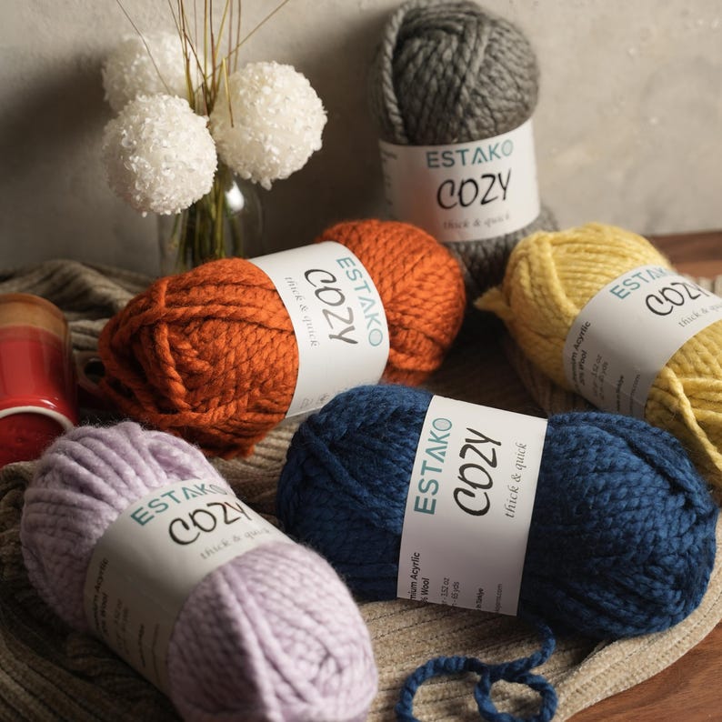 Pode incluir: Uma cole&ccedil;&atilde;o de novelos de l&atilde; coloridos em tons de laranja, cinza, amarelo, azul e lavanda. Cada novelo tem uma etiqueta que diz "ESTAKO COZY thick & quick". A l&atilde; est&aacute; disposta sobre uma superf&iacute;cie de tons neutros, com esferas brancas decorativas e um vaso no fundo.