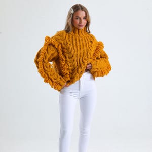 Pode incluir: Camisola de malha grossa amarelo mostarda com gola alta e mangas com babados. A camisola apresenta um design de malha entran&ccedil;ada na frente e nas mangas. A modelo est&aacute; a usar cal&ccedil;as de ganga brancas. &Eacute; uma pe&ccedil;a de roupa quente e aconchegante.