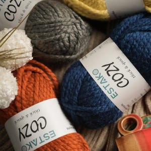 Pode incluir: Close-up de v&aacute;rios novelos de l&atilde; grossa e grossa em v&aacute;rias cores, incluindo cinza, laranja e azul marinho. Cada novelo tem um r&oacute;tulo que diz "ESTAKO COZY thick & quick". Ideal para projetos de tric&ocirc; ou croch&ecirc;.