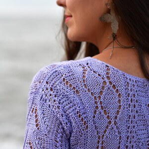 May include: A light purple, short-sleeved knit top with a delicate openwork pattern. The top has a scoop neckline and scalloped edges on the sleeves. The knitwear is a soft, pastel color.