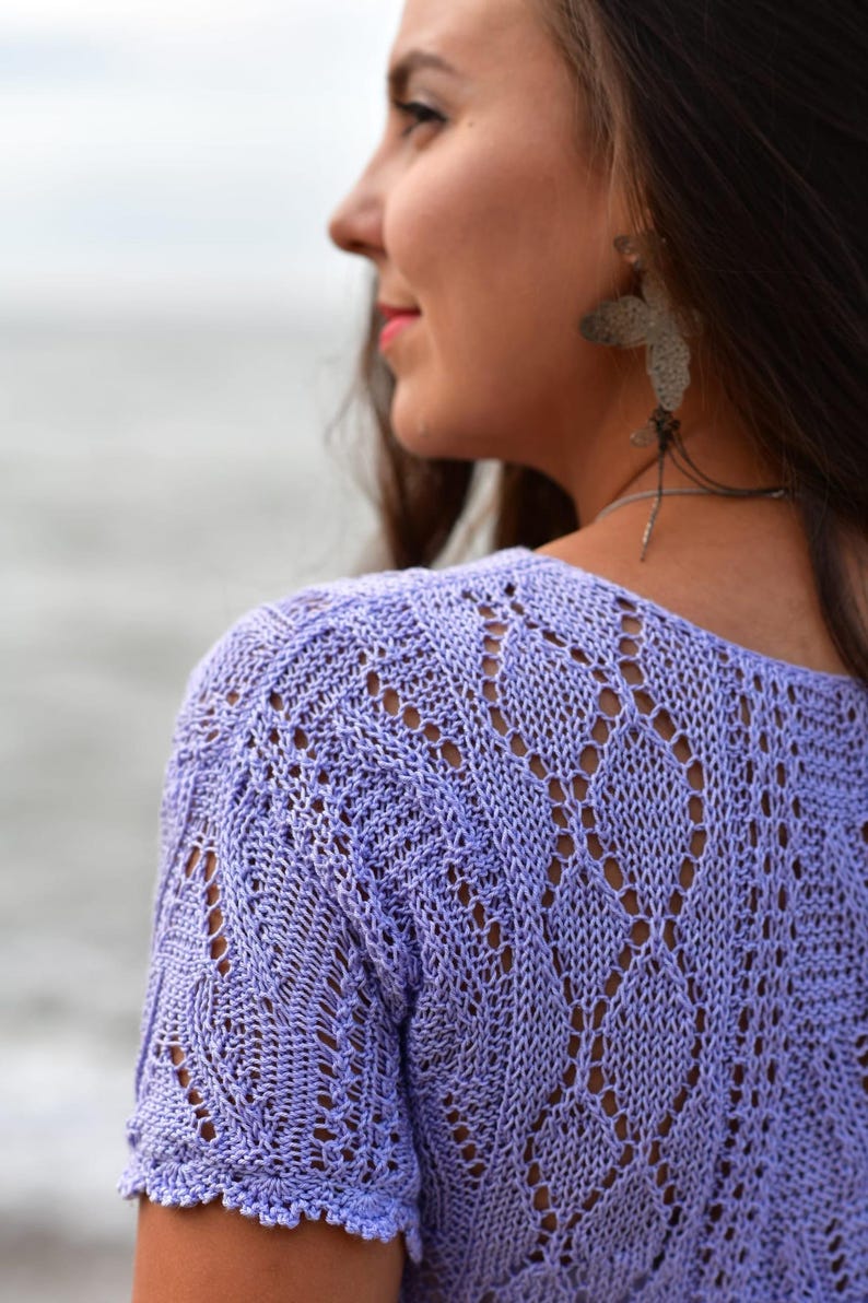 May include: A light purple, short-sleeved knit top with a delicate openwork pattern. The top has a scoop neckline and scalloped edges on the sleeves. The knitwear is a soft, pastel color.