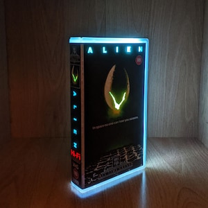 May include: Illuminated VHS tape of the movie "Alien" with a glowing blue frame. The cover art features a glowing egg and the film's title. The text "In space no one can hear you scream" is also visible.