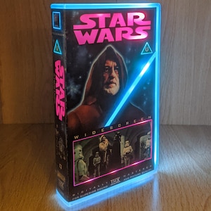 May include: A Star Wars VHS tape converted into a light-up display. The box features the movie title in pink, an image of Obi-Wan Kenobi, and a blue LED frame. The words "Widescreen" and "Digitally Mastered" are also visible.