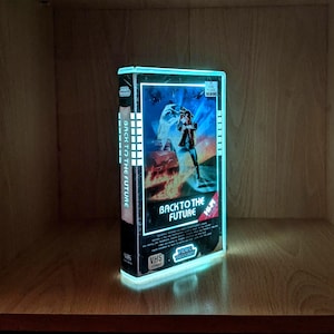 May include: A lit-up VHS tape of the film "Back to the Future" on a wooden shelf. The tape case has a blue neon glow. The movie title is visible, along with the VHS format logo. The overall effect is a retro, glowing display.