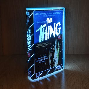 Könnte beinhalten: Eine leuchtende VHS-Kassette des Films "The Thing" mit blau-weißer Neonbeleuchtung. Auf dem Cover der Kassette sind der Filmtitel und der Slogan "Man is the warmest place to hide" zu sehen.