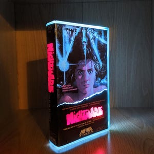 May include: A lit-up VHS tape of the film "Nightmare" with a blue and red light border. The movie title is in pink, with the film's title and actors' names visible. The image features a woman with a terrified expression.