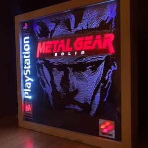 May include: A rectangular, white-framed light box featuring the Metal Gear Solid video game cover art. The display shows the title in red, a blue-toned character portrait, the PlayStation logo, an ESRB M rating, and the Konami logo in the corner.