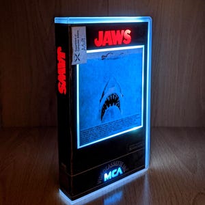 May include: A lit-up VHS tape of the film "Jaws." The black case features the title in red and a blue shark image. The tape is labelled "Video Cassette Inc MCA" and has a "Department of Consumer Affairs Adult" sticker.