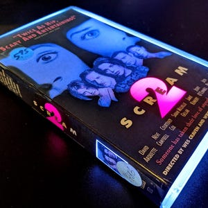Puede incluir: Una cinta VHS de la película "Scream 2" con fondo negro. La portada muestra el título en rosa y caras de actores. La cinta tiene el texto "Twice As Hip, Scary And Entertaining!"