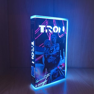 Puede incluir: Una cinta VHS de Disney TRON iluminada con un borde de luz LED azul brillante. La portada presenta el título TRON en letras blancas sobre ilustraciones de personajes con trajes brillantes sobre un fondo oscuro con líneas rosas.