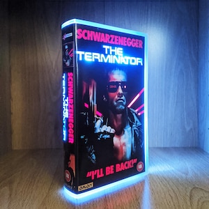 May include: A glowing VHS tape of "The Terminator" starring Arnold Schwarzenegger. The box art is predominantly black, with the title in blue and the quote "I'll be back!" in red. The edges of the tape are highlighted with a blue light.