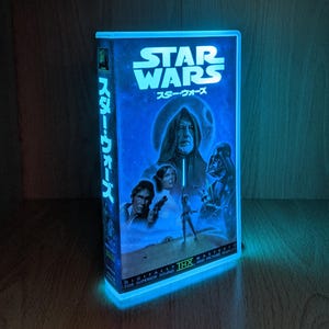 Può includere: Una luce notturna a forma di cassetta VHS di Star Wars blu brillante. La parte anteriore presenta il logo di Star Wars e testo giapponese. L'immagine include Darth Vader, Luke Skywalker, la Principessa Leia e Han Solo. La parte inferiore del nastro recita "DIGITALLY THX MASTERED FOR SUPERIOR SOUND AND PICTURE QUALITY."